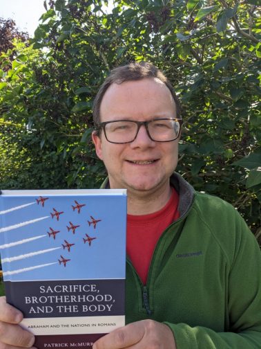 Dr McMurray holding a copy of his book, Sacrifice, Brotherhood, and the Body: Abraham and the Nations in Romans. The cover has an image of airplanes flying in formation, which is symbol or metaphor to represent the nations and Israel being brought into the correct alignment or formation.
